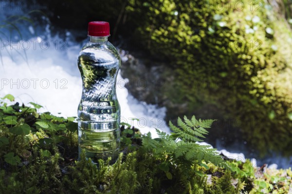 Transparent plastic A bottle of clean water with a red lid stands in the grass and moss on the background of a rugged mountain river. The concept of pure natural drinking water