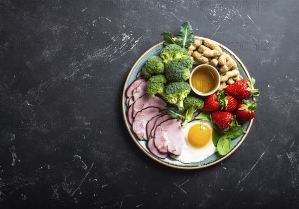 Ketogenic low carbs diet, top view, space for text. Plate on stone black background with keto foods: egg, meat, olive oil, broccoli, berries, nuts, seeds. Healthy fats, clean eating for weight loss