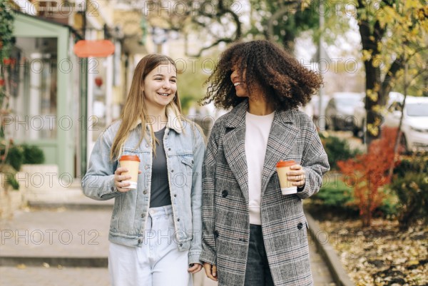 Two happy carefree multiracial girlfriends walking city street with take away coffee, talking and sharing life stories, multiethnic female best friends enjoying autumn walk outdoors. Womens friendship