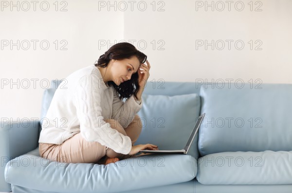 Young pensive woman in casual wear sitting with her legs on sofa using keyboard on laptop, staring at computer screen, looking for important necessary information or answers urgent emails