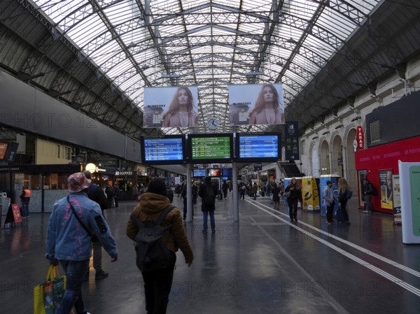 Travellers in a station concourse under a large glass-covered ceiling with timetable displays and advertising, Gare le Est .Paris