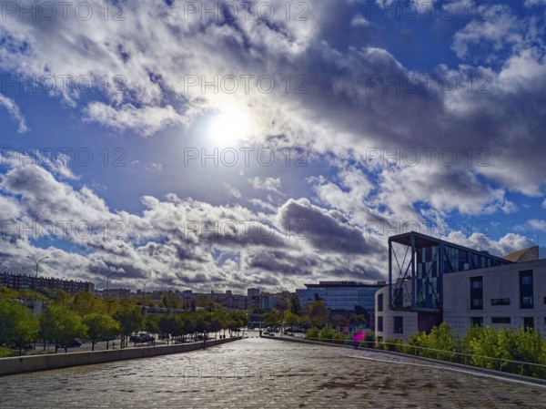 The sun shines through the clouds over an urban landscape with people, trees and modern buildings, forecourt of the Philharmonie de Paris. Paris