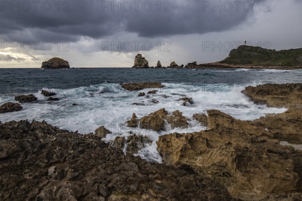 A rocky coastline with a pointed rock formation stretches into the sea at a bay, strong waves at a rainy sunrise create a dramatic atmosphere. Pointe des Chateau with view of Pointes des colibris on Grande Terre, Guadeloupe, French Antilles, Caribbean, North America