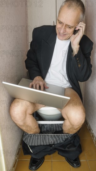 A workaholic adult businessman is using the computer laptop and the mobile phone, also called smartphone, while he is sitting on the bowl in the toilet
