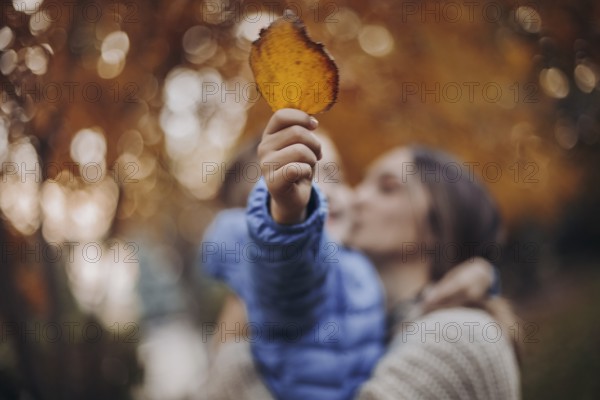 Young happy family, mother and little son child walking in autumn forest, kid holding yellow tree leaf in hand while spending time with mom in beautiful fall nature