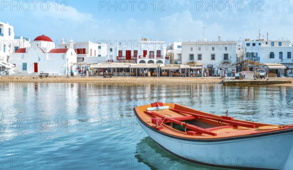 Beautiful summer day sunshine on Greek island. Fishing boats moored at jetty. Whitewashed houses by waterfront. Red dome church, restaurants, shops, fish market stalls. Mediterranean lifestyle. Small boat in foreground. Mykonos, Cyclades, Greece, Europe