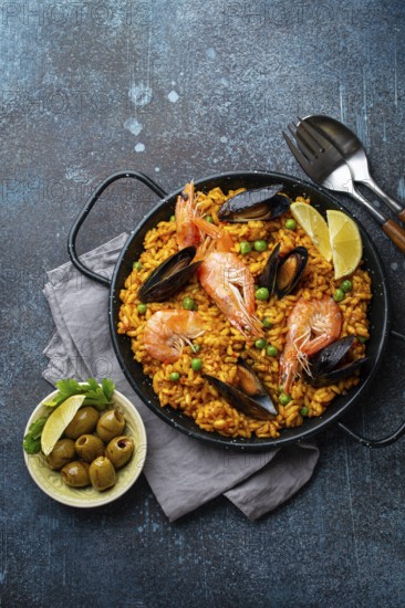 Classic dish of Spain, seafood paella in traditional pan on rustic blue concrete background top view. Spanish paella with shrimps, clamps, mussels, green peas and fresh lemon wedges from above