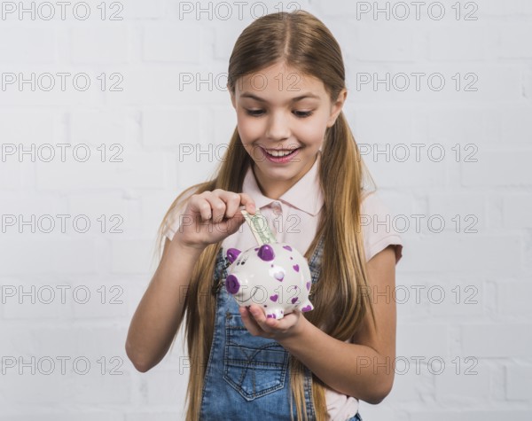 Smiling portrait girl inserting currency note white piggybank