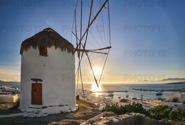 Romantic view of traditional Greek whitewashed windmill on hill. Chora town and harbor of Mykonos, Greece, low sun above sea horizon, sunset sky. Vacations, leisure, Mediterranean lifestyle, Europe