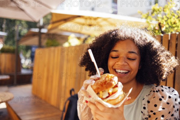 Young african american girl eating classic burger and drinking lemonade in the city. Outdoor woman wearing summer blouse with polka dots eatting delicious burger while sitting at terrace. Woman in blue tasting sandwich having joyful look admiring sunny w