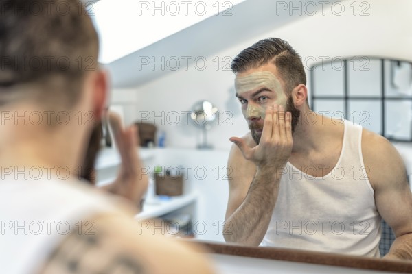 Reflection in the bathroom mirror of a young bearded man applying a face mask to his skin in a skincare and hygiene concept