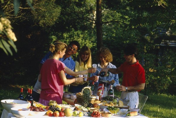 Garden party, food and drink, richly laid table, fruit, vegetables, fruits, bread, cheese, wine plates, bowls, young people, 3 woman, 2 men, 5 people, toast, picnic, eating outdoors