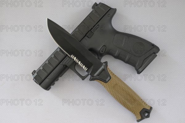 A hunting knife with a beige handle lies on a black pistol on a white surface