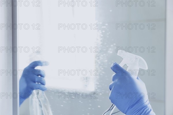 Cropped view of housekeeper woman hand in rubber blue gloves spraying chemical cleaner liquid to the mirror while cleaning bathroom at home. Housekeeping and cleaning service. Clean house, cleanliness