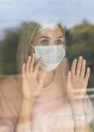 Front view woman with medical mask home touching window during pandemic