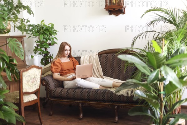 Barefoot female using netbook while relaxing on elegant couch amidst lush potted plants