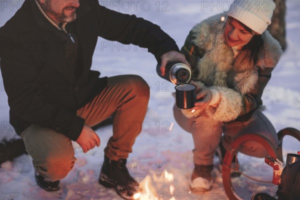 Joyful happy family couple in love basking by campfire outside in winter snowy forest drinking hot tea together, man and woman warm themselves hugging and loving look at each other. Trekking, adventure and seasonal vacation