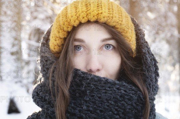 Portrait of a smiling young caucasian woman in a bright woolen hat and scarf, against the backdrop of a snowy forest