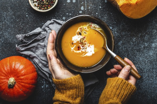 Woman in yellow knitted sweater eating a pumpkin cream soup on dark stone background with spoon decorated with cut fresh pumpkin, top view. Autumn cozy dinner concept