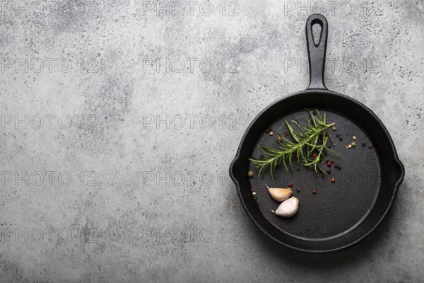 Black cast iron frying pan with ingredients for cooking: fresh green rosemary, raw garlic cloves and colorful peppercorns on gray stone background flat lay top view with space for text