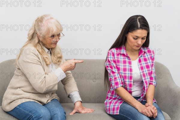 Senior mother scolding her adult daughter sitting sofa against wall
