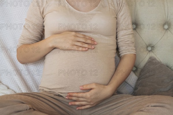 Cropped shot of pregnant woman holding belly while relaxing on couch at home, expectant mother sitting on sofa indoors and gently touching tummy. Pregnancy, maternity and prenatal period concept