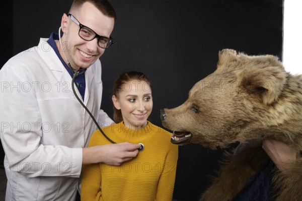Smiling young doctor with stethoscope and patient examining bear, studio medical humor of whom to treat, human or animal