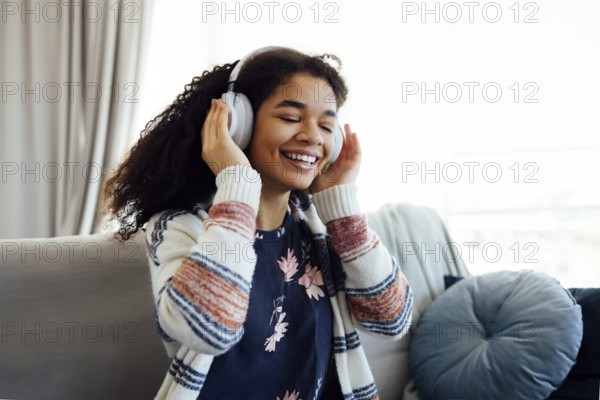 African millennial woman in orange sweater wearing white wireless headphones, listening music, resting and lying on couch in living room. Girl spend lazy weekend at home