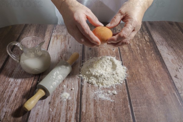 Woman breaking an egg, pouring it over flour to make a sponge cake on a wooden table with a rolling pin and a pitcher of milk