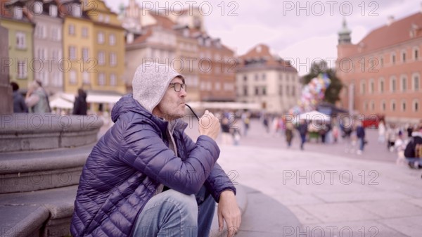 Portrait of adult man with glasses sitting in the hood on square and smoking a tobacco pipe in the Palace Square, Warsaw Old Town