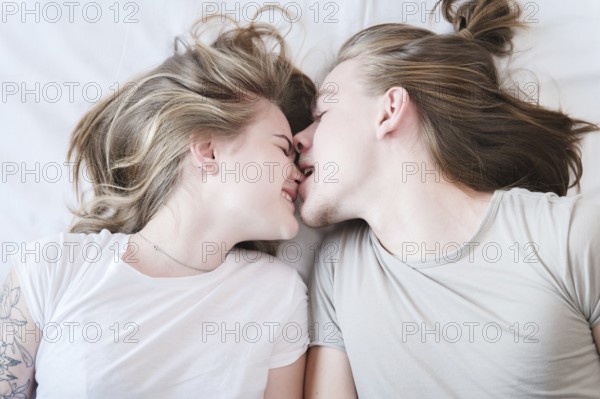 A top view of a molo pair of long-haired men and women laughing around fooling around in bed. A man bites a girl by the nose. The concept of a happy young family