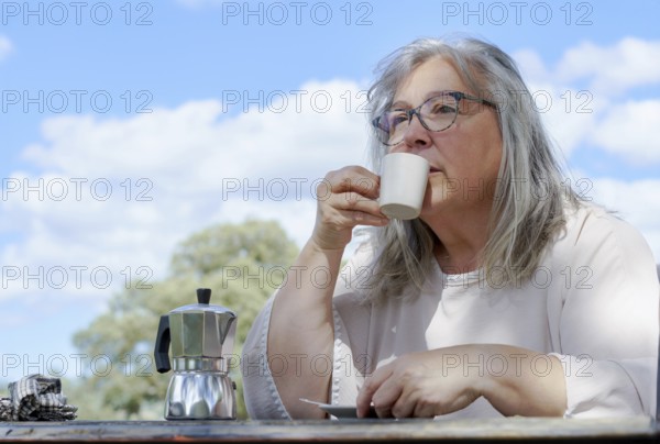Close-up of an older woman with white hair drinking a cup of coffee at a table in a field with a cloudy blue sky in the background