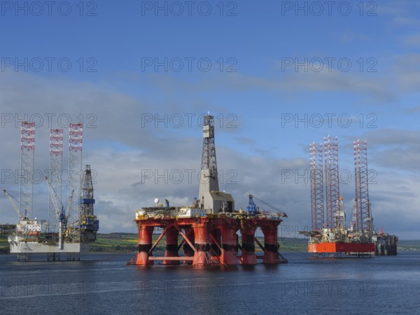 Three oil rigs standing side by side in a calm sea under a blue sky, partly cloudy, drilling rigs in red against a blue sky in the north sea, inverness, Scotland, United Kingdom, Europe