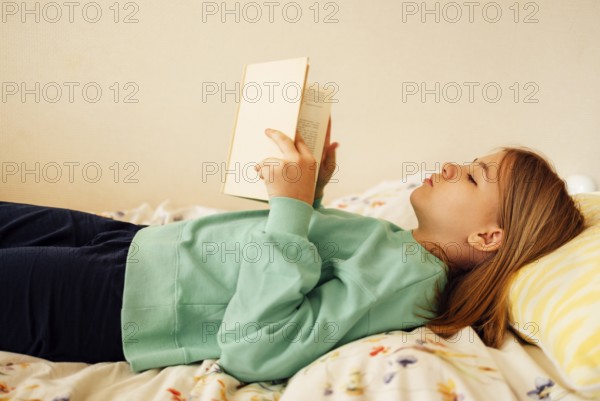 Caucasian blond teenage girl is lying on the bed and reading book. Reading fiction, hobbies, leisure activities and recreation. Concept of home education