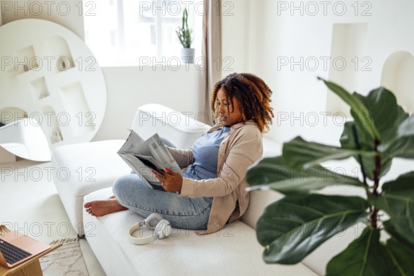 Photo of pretty happy young african woman sitting on sofa indoors at home while reading magazine or book