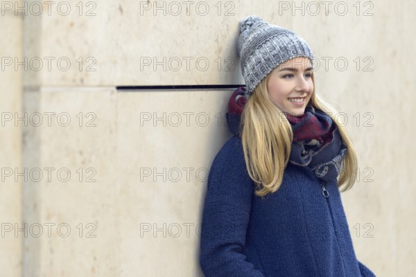 Smiling attractive young blond woman in a warm blue winter outfit standing daydreaming leaning back against an exterior wall staring into the distance