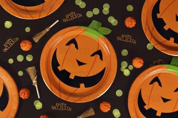 Halloween party flat lay with orange pumpkin paper plates, witch brooms, confetti and Happy Halloween text on black background