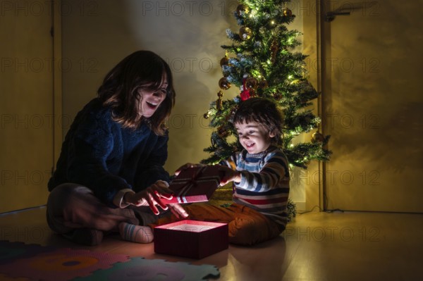 A woman and a child are opening a present in front of a Christmas tree. Scene is joyful and festive, as the two are sharing a special moment together during the holiday season