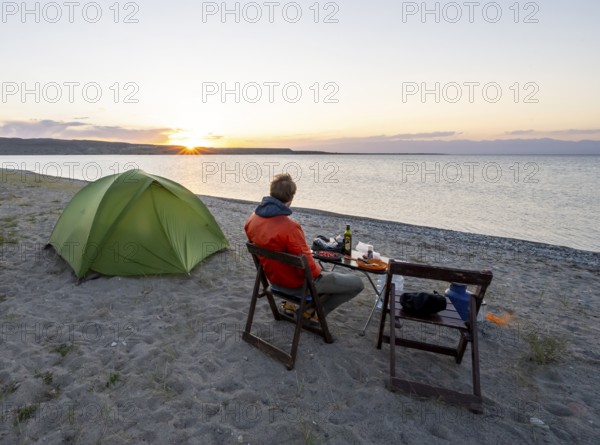 Camping at the lakeshore, young man sitting next to tent on camping chair during dinner, sunset at Issyk Kul Lake, Issyk Kul, Kyrgyzstan, Asia