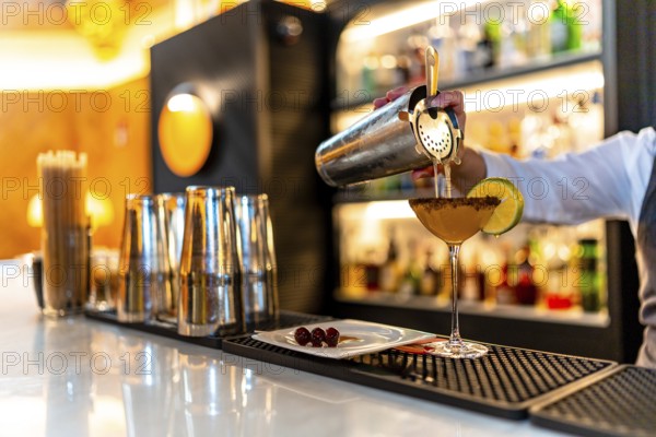 Hand of a waiter filling cocktail into martini glass garnished with lemon in the counter of a luxury night club