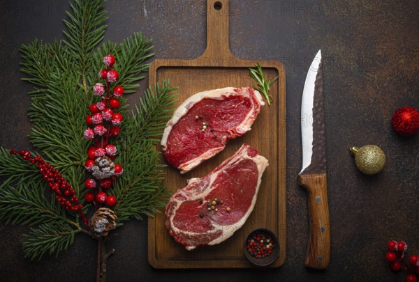 Two raw uncooked meat beef steaks on wooden cutting board with knife and seasonings on dark rustic background with Christmas festive decoration from above, food photography
