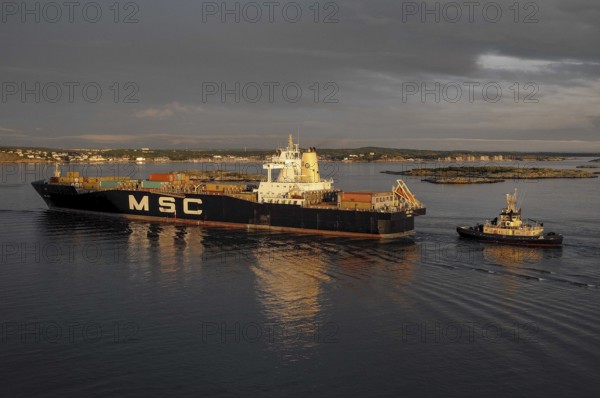A cargo ship and a tugboat sail on calm water at sunset, loaded with containers, Gothenburg, Baltic Sea, Sweden, Europe