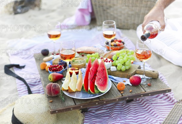 Crop male serving pink wine having picnic with delicious snacks and fruit on sandy beach