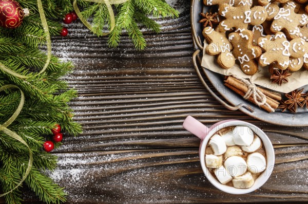 Flat lay Christmas background of pink mug with hot chocolate and marshmallows, spruce branch and tray with gingerbread man cookies on wooden table