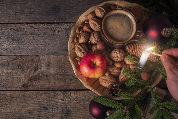 Christmas table with a wooden plate full with nuts, apples, a cup of coffee and green fir twigs, while a man lights up a candle. Low light image