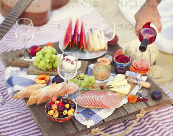 Crop male serving pink wine having picnic with delicious snacks and fruit on sandy beach