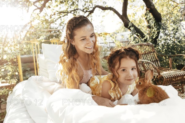 Young woman and cute little girl with fluffy rabbit relaxing on bed on sunny summer day in garden