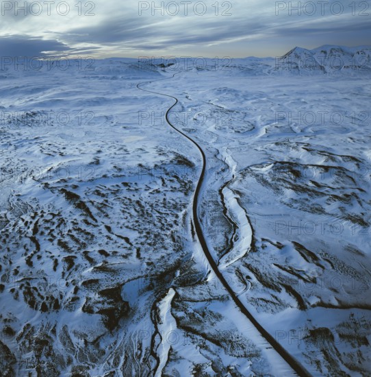 Aerial view of a long road winding through a vast snowy landscape with distant mountains under a cloudy sky, North, Iceland, Europe