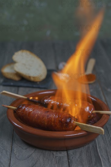 Typical spanish tapa, chorizo on fire in an earthenware casserole with slices of bread in the background