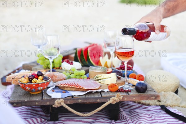 Crop male serving pink wine having picnic with delicious snacks and fruit on sandy beach